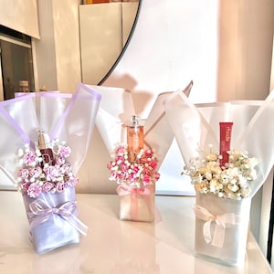 以下が含まれることがあります： 香水瓶とリップ製品が入った3つのギフトブーケ。それぞれ白とパステルカラーの紙で包まれ、リボンで結ばれ、小さな花で飾られています。ブーケは反射する表面に配置されています。