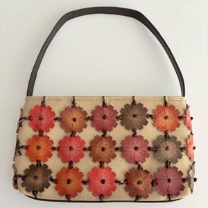 May include: A beige canvas handbag featuring a grid of leather flower cutouts in shades of red, orange, and brown. The flowers are attached with dark brown cord knots. The bag has a single dark brown leather shoulder strap and a rectangular shape.