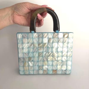 May include: A square handbag constructed from a grid of iridescent, square-shaped mother-of-pearl tiles linked together with light blue thread. The bag features a single, rounded, dark brown, semi-translucent handle attached to the top edge.