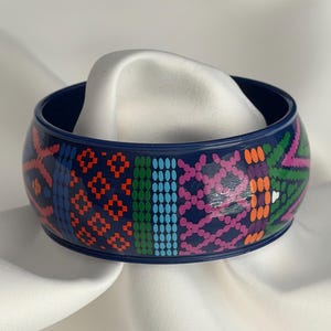 May include: A dark blue bangle bracelet featuring a repeating pattern of geometric shapes and dots in vibrant colors including orange, light blue, green, and pink. The surface of the bracelet is smooth and glossy, showcasing a multi-colored design.