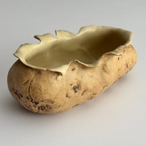 May include: A handmade ceramic bowl with a unique, organic shape. The exterior has a textured, earthy brown appearance with dark spots, while the interior and rim are a lighter, creamy beige. The rim has a ruffled, leaf-like design.