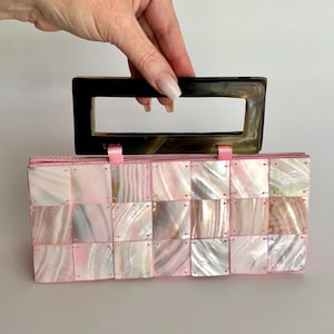 May include: A rectangular pink and white mother-of-pearl handbag with a dark brown handle. The bag is made of small, square iridescent tiles. Pink ribbon accents are visible on the top of the bag.
