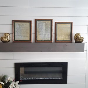 Op de afbeelding: Drie ingelijste historische documenten met bruine houten frames worden boven een zwarte open haard weergegeven. Twee gouden hartvormige decoratieve objecten staan aan weerszijden van een grijs houten plank. De documenten lijken verouderd.