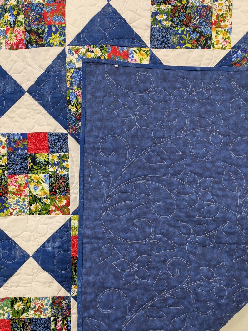 May include: A handmade quilt with a patchwork design featuring squares of floral fabric in red, blue, and white. The quilt has a blue and white diamond pattern and a solid blue backing with floral embroidery.