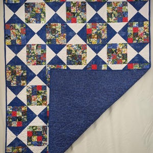 May include: A handmade quilt with a patchwork design. The quilt features squares of floral fabric in red, yellow, and blue, set against white and blue geometric shapes. The reverse side is a solid blue with a floral pattern.