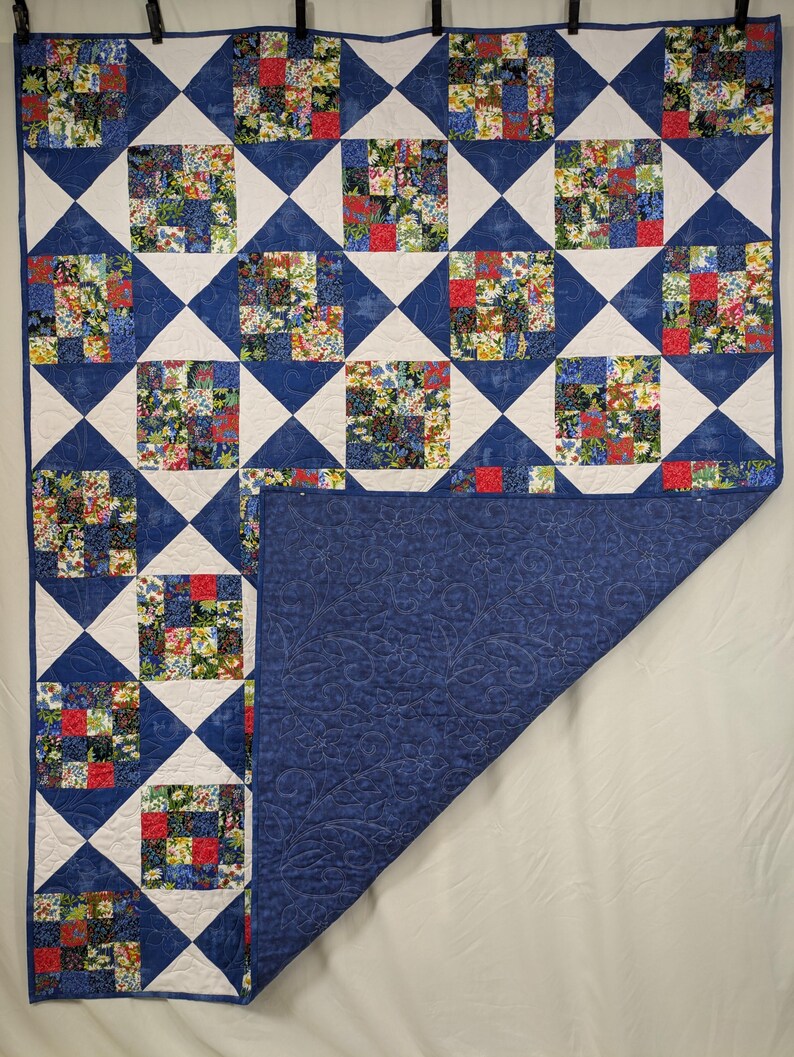 May include: A handmade quilt with a patchwork design. The quilt features squares of floral fabric in red, yellow, and blue, set against white and blue geometric shapes. The reverse side is a solid blue with a floral pattern.