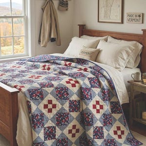 May include: A patchwork quilt with a blue, red, and cream color scheme covers a wooden bed. The quilt features a repeating pattern of squares with floral and cross designs. A small pillow with the word "VERMONT" is on the bed.