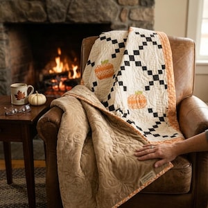 May include: A reversible quilt featuring a cream-colored side with a black grid pattern and orange pumpkin motifs, and a solid tan side with embossed pumpkin designs. The quilt has an orange patterned border and a small rectangular label.