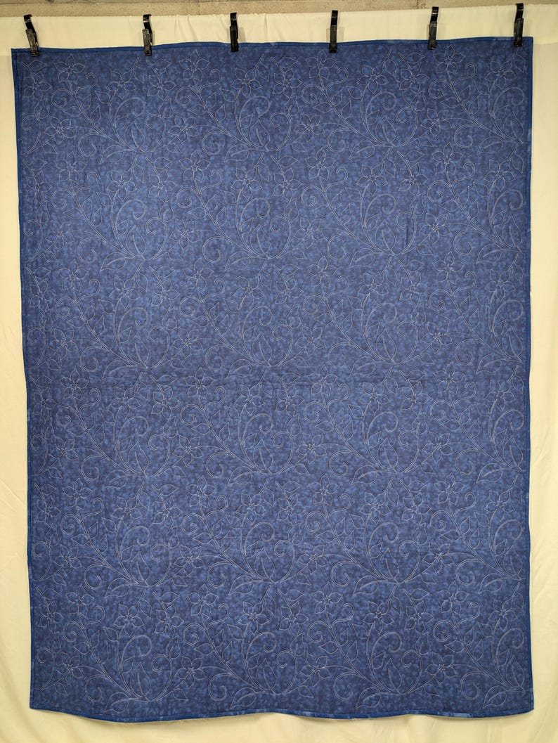 May include: A blue quilt with a floral pattern. The quilt is a solid blue color with a detailed, repeating floral design stitched throughout. The quilt is hanging from black clips.