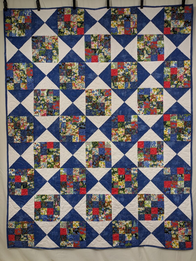 May include: A handmade quilt with a repeating pattern of squares and diamonds. The quilt features a combination of floral fabric squares in red, yellow, and blue, set against a white and blue background. The quilt is a cozy and decorative textile.