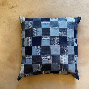 Pode incluir: Almofada quadrada de ganga com design de patchwork. A almofada apresenta um padrão de grade de quadrados de ganga azul de várias tonalidades, criando uma superfície texturizada e visualmente interessante. Um elemento decorativo para a casa.