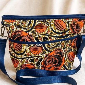 May include: A rectangular crossbody bag with a vibrant floral pattern in orange, black, and olive green. The bag has a navy blue trim, a matching adjustable strap, and metal hardware. The bag is approximately 10 inches wide.