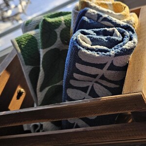 May include: A wooden crate with a green knob holds rolled-up towels. The towels are patterned with leaves in green, blue, and yellow. The crate is made of light brown wood.