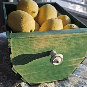 May include: A green wooden box filled with yellow lemons. The box has a white and black knob. The lemons are a bright yellow colour and appear fresh. The box is on a white lace tablecloth. The scene is lit by natural light.