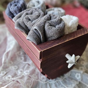 Puede incluir: Una caja de almacenamiento de madera de color burdeos con un asa en forma de estrella, llena de toallas enrolladas y esponjosas en tonos grises y blancos. La caja está sobre una superficie de encaje blanco.