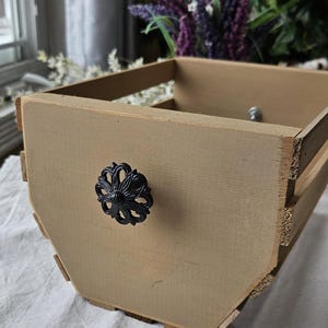 May include: A light brown wooden crate with a black floral knob. The crate has a slatted design and is painted a neutral color. The knob is centered on the front panel. The crate is likely used for storage or decoration.