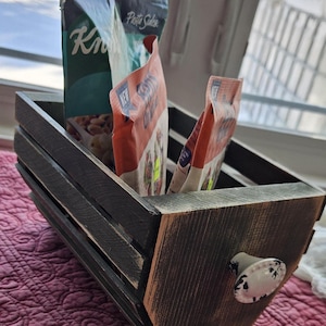 Puede incluir: Una caja de madera rústica con un asa decorativa, que contiene varios paquetes de alimentos. La caja está teñida de marrón oscuro con vetas de madera visibles y un asa con estampado en blanco y negro. Los paquetes incluyen "Pasta Sides" y "Knorr".