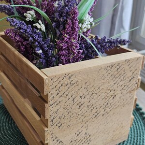 May include: A wooden crate filled with purple and white faux lavender and green leaves. The crate has a light brown finish with script writing on the front. The crate sits on a green placemat.