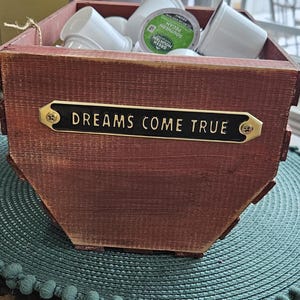Peut inclure: Une boîte en bois rustique, de couleur marron foncé, contenant des dosettes de café blanches et une dosette verte. Une plaque noire avec l'inscription dorée "DREAMS COME TRUE" est fixée à l'avant de la boîte.