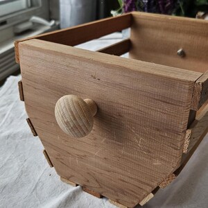 Peut inclure: Une boîte en bois faite à la main avec un bouton en bois arrondi. La boîte est construite en bois brun clair avec un grain visible et est rectangulaire avec un devant incurvé. La boîte a une esthétique rustique et champêtre.