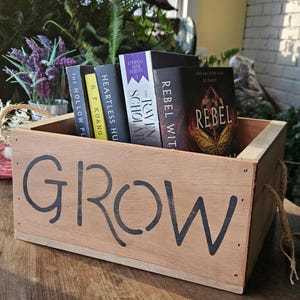 Peut inclure: Une caisse en bois avec le mot "GROW" peint en noir. La caisse contient plusieurs livres, dont "REBEL WIT" et "THE RAVEN SCHOLAR". La caisse a des poignées en corde et est posée sur une surface en bois.