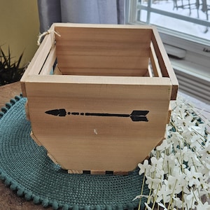 Peut inclure: Une caisse en bois brun clair avec un motif de flèche noire. La caisse est rectangulaire avec un design à lattes et une poignée en corde. Elle repose sur un dessous-de-plat turquoise avec des fleurs blanches à proximité.