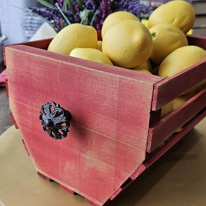 May include: A rustic red wooden crate filled with yellow lemons. The crate has a decorative black handle and is set on a gold-colored surface. Purple flowers and greenery are in the background.