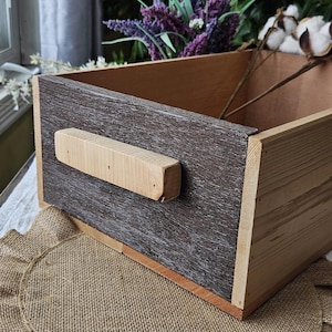 Peut inclure: Une boîte de rangement en bois avec une finition en bois naturel et un panneau avant texturé gris foncé. Une poignée en bois de couleur claire est fixée à l'avant. La boîte est vide et semble faite à la main.