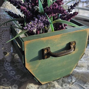 Op de afbeelding: Een rustieke, groene houten plantenbak met een verweerde afwerking, gevuld met levendige paarse kunstlavendel en groen blad. De bak heeft een metalen handvat en staat op een kanten tafelkleed, wat een charmante, boerderij-esthetiek creëert.