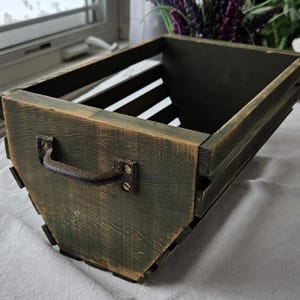 Peut inclure: Une caisse en bois rectangulaire avec une finition vert foncé et une poignée en métal rustique. La caisse a un design à lattes sur les côtés et est construite avec un grain de bois visible. La poignée est fixée avec des vis de couleur laiton.
