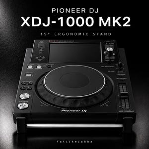 Op de afbeelding: Een zwarte Pioneer DJ XDJ-1000 MK2 digitale mediaspeler gemonteerd op een ergonomische standaard van 15 graden. Het apparaat is voorzien van een groot centraal jogwheel, een touchscreen aan de bovenzijde, diverse bedieningsknoppen en een zilveren horizontale basis.
