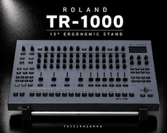 Könnte beinhalten: Eine graue Roland TR-1000 Rhythm Creator Drum-Machine auf einem ergonomischen 15-Grad-Ständer. Das Gerät verfügt über zahlreiche schwarze Regler, Schieberegler und Tasten auf dem Bedienfeld, mit dem Text ROLAND TR-1000 15° ERGONOMIC STAND.