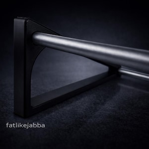 Puede incluir: Un soporte triangular de metal negro que sostiene una varilla cil&iacute;ndrica plateada horizontal. El soporte presenta un dise&ntilde;o de marco s&oacute;lido con un acabado mate suave. El texto fatlikejabba est&aacute; impreso en blanco abajo a la izquierda.