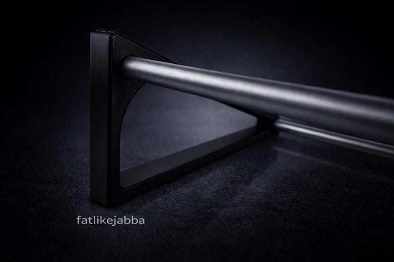 Puede incluir: Un soporte triangular de metal negro que sostiene una varilla cil&iacute;ndrica plateada horizontal. El soporte presenta un dise&ntilde;o de marco s&oacute;lido con un acabado mate suave. El texto fatlikejabba est&aacute; impreso en blanco abajo a la izquierda.