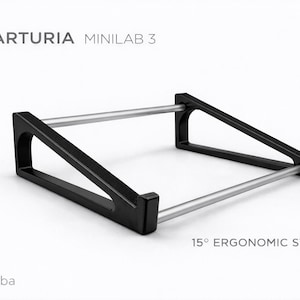 May include: Black ergonomic stand for Arturia Minilab 3, with a 15-degree angle. The stand features a rectangular frame with two silver bars and black angled supports. The text "ARTURIA MINILAB 3" and "15° ERGONOMIC STAND" are visible.