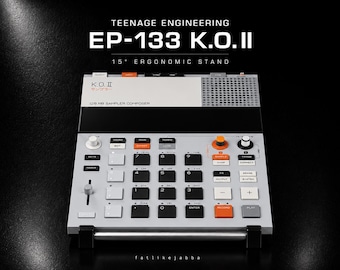 Könnte beinhalten: Ein Teenage Engineering EP-133 K.O. II Sampler-Composer auf einem 15-Grad-Ergonomieständer. Das Gerät verfügt über ein hellgraues Gehäuse, ein Raster aus schwarzen und weißen Tasten, orangefarbene Akzenttasten, einen Fader und ein Lautsprechergitter.