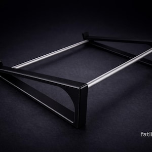 Puede incluir: Un soporte de metal negro con dos marcos laterales triangulares conectados por dos varillas met&aacute;licas horizontales de color plateado. El soporte tiene un dise&ntilde;o angular y elegante con un acabado mate. El texto fatlikejabba est&aacute; impreso en blanco.