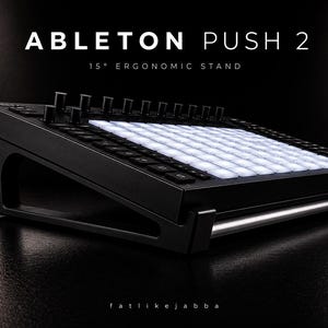 Puede incluir: Controlador de producción musical Ableton Push 2 negro sobre un soporte ergonómico de 15 grados. El controlador tiene una cuadrícula de pads blancos iluminados, botones negros y detalles plateados. El texto "ABLETON PUSH 2" está en la parte superior.