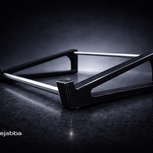 Puede incluir: Un soporte negro para port&aacute;til con dos soportes laterales triangulares conectados por dos varillas met&aacute;licas horizontales plateadas. El soporte tiene un dise&ntilde;o de perfil bajo con peque&ntilde;os ganchos frontales. El texto fatlikejabba est&aacute; impreso en blanco.