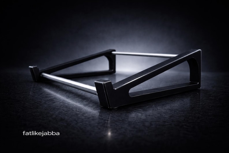 Puede incluir: Un soporte negro para port&aacute;til con dos soportes laterales triangulares conectados por dos varillas met&aacute;licas horizontales plateadas. El soporte tiene un dise&ntilde;o de perfil bajo con peque&ntilde;os ganchos frontales. El texto fatlikejabba est&aacute; impreso en blanco.
