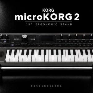 Puede incluir: Un sintetizador Korg microKORG 2 de color gris oscuro sobre un soporte ergonómico de 15 grados. El teclado cuenta con teclas blancas y negras, una pantalla central, varios mandos de control y botones. El texto KORG microKORG 2 15° ERGONOMIC STAND es visible arriba.