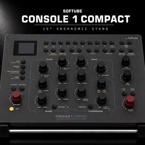 May include: A black Softube Console 1 Compact channel strip control system featuring a 15-degree ergonomic stand. The device has numerous black knobs, two red knobs, a small display screen, and various buttons labeled with white and orange text.
