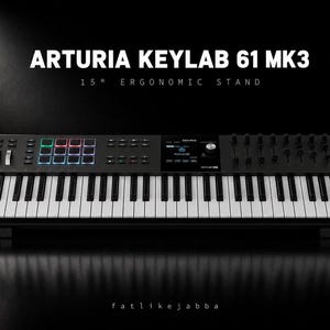 May include: Arturia Keylab 61 MK3 keyboard controller with a 15 degree ergonomic stand. The black device features white and black keys, a central display screen, multi-colored backlit pads, and wooden side panels on a dark background.