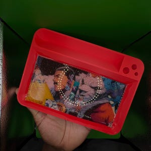 May include: A red rectangular tray with a colorful image of a person with a mustache and other figures. The tray has a small compartment and three circular holes. The image is inside the tray.