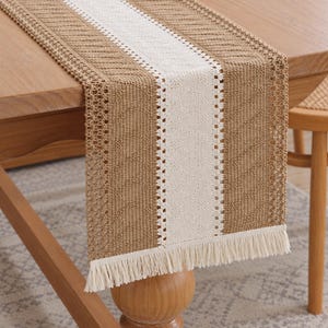 Puede incluir: Un camino de mesa rectangular con un panel central de encaje color crema flanqueado por dos secciones más anchas de tela de ganchillo color canela. El camino de mesa termina con un fleco corto y recto de hilo color crema en el borde.