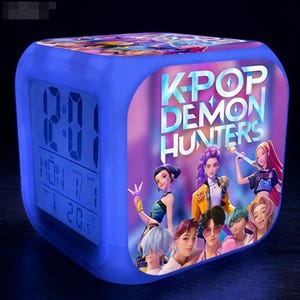 May include: A blue cube-shaped digital alarm clock with the text "K-POP DEMON HUNTERS" and images of animated characters. The clock displays the time, day, date, and temperature.