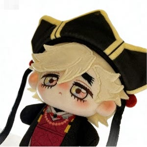 May include: A plush doll with light blonde hair and embroidered facial features, wearing a black hat with gold trim and a red and black outfit. The doll has black ribbons and red accents.