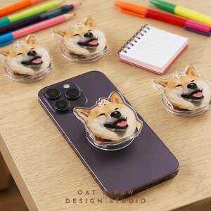 May include: A purple smartphone with a clear phone grip featuring a smiling Shiba Inu dog design. The image includes three additional matching grips, pens, and a small notepad. The text "OAT & PAW DESIGN STUDIO" is visible.