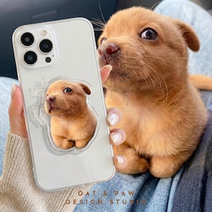 Può includere: Custodia trasparente per telefono con un design a cucciolo. La custodia ha un retro trasparente e un'immagine in rilievo di un piccolo cucciolo marrone. Il cucciolo nell'immagine ha un mantello marrone chiaro e guarda in alto. La custodia del telefono è tenuta in mano da una persona.