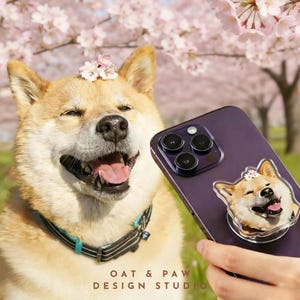 May include: A smiling Shiba Inu dog with pink flowers on its head, next to a purple phone with a matching dog phone grip. The phone grip features a smiling Shiba Inu with a flower crown. The text "OAT & PAW DESIGN STUDIO" is at the bottom.